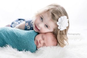 texas roots a newborn boy in a Milwaukee photography studio wrapped in a blue swaddle is laying on top of white fur rug his older sister who is three has her head above newborn baby and is smiling at the camera backlighting in image makes for a bright white backdrop