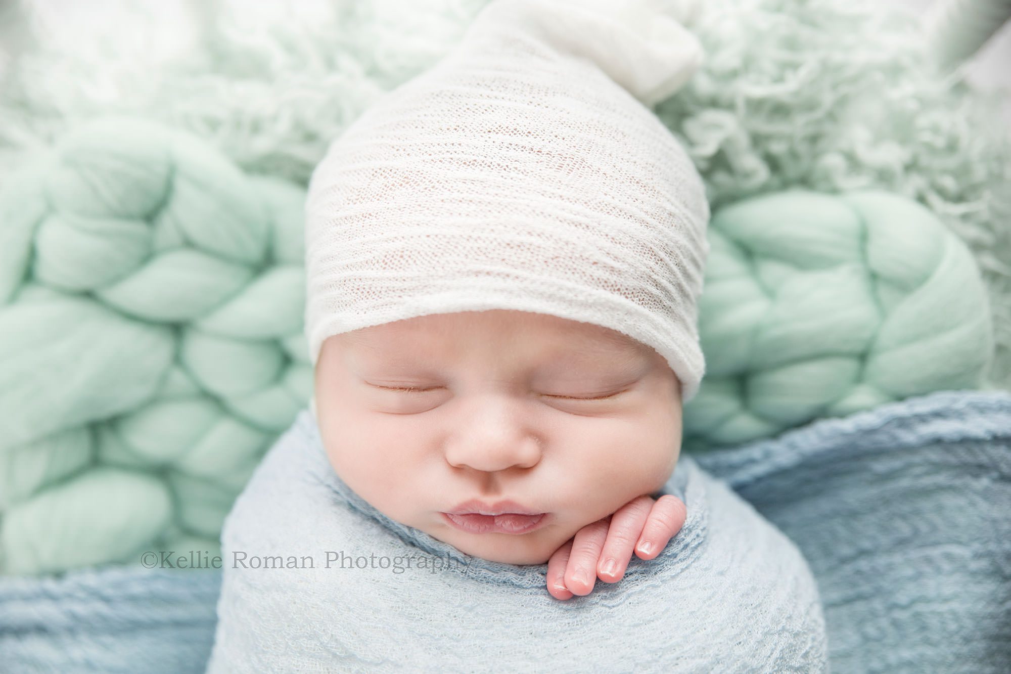 texas roots a newborn boy from kenosha in a Milwaukee photographers studio he is sleeping onto of a mint colored layering fabric with a white hat on and a blue wrap over him his left fingers are sticking out of the wrap