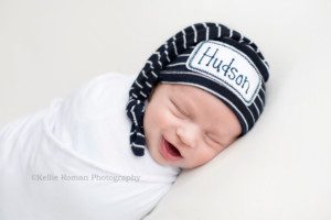 texas roots a newborn boy from kenosha in a Milwaukee photography studio baby is wrapped in a white wrap onto of an ivory blanket boy has a striped hat on that says his name he has his eyes closed and is smiling very big