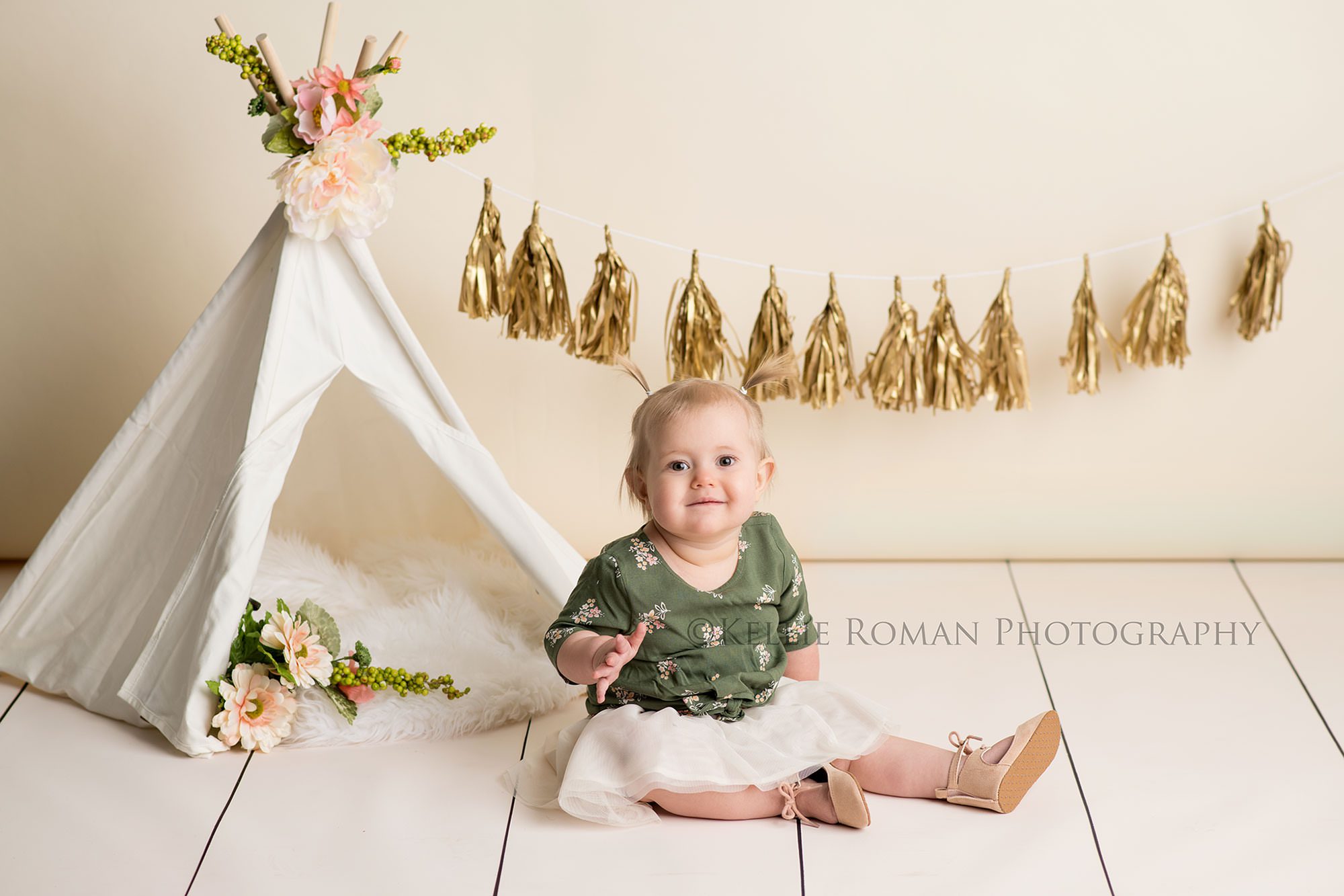 floral milestone session one year old girl from kenosha Wisconsin in a Milwaukee Wisconsin photographer studio sitting on a wood floor with a cream backdrop behind her She's sitting next to a small teepee with flowers inside of it and a gold tassel garland little girl has on a green floral top with a tutu skirt