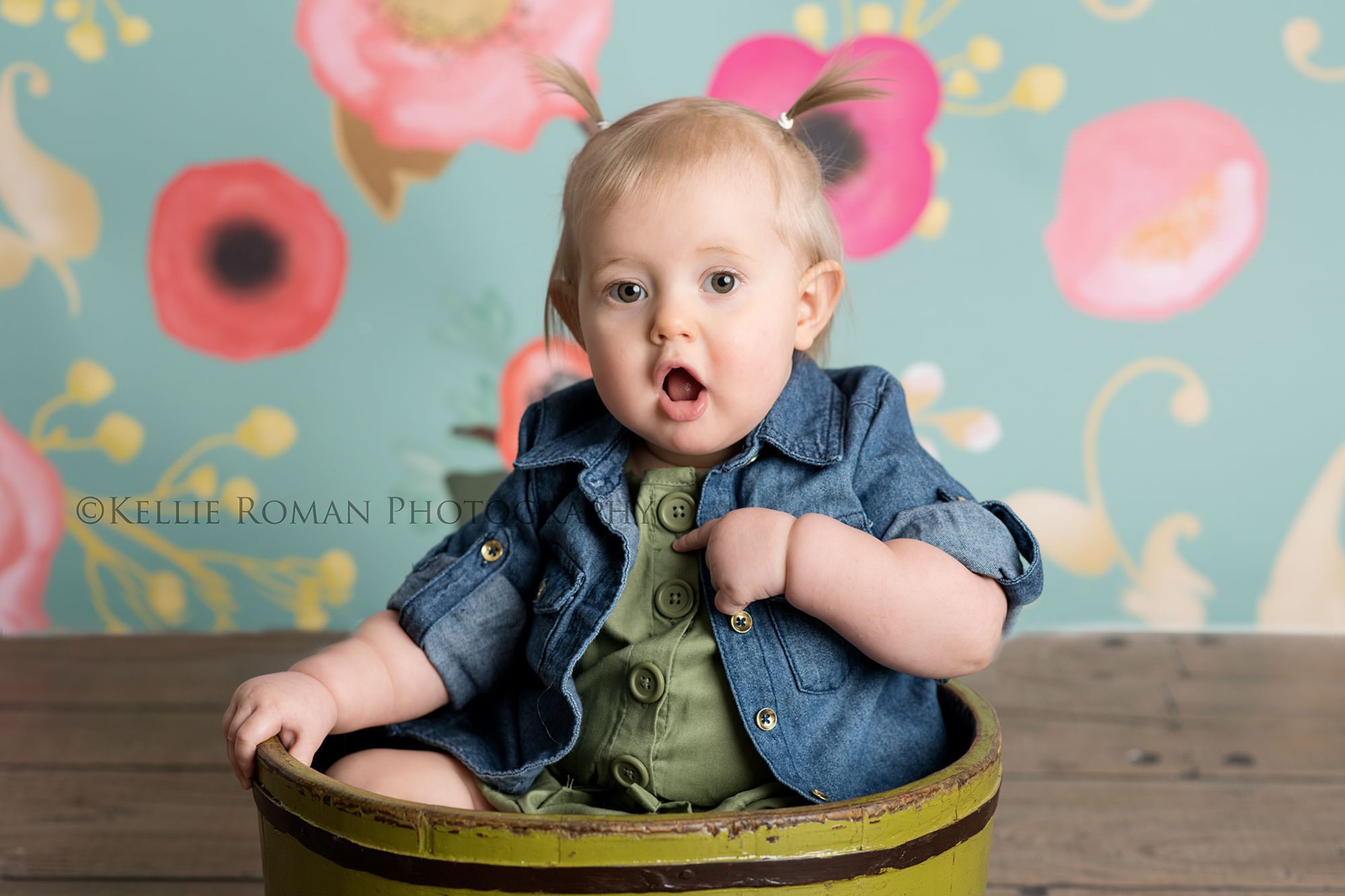 floral milestone session one year old girl from kenosha in a Milwaukee Wisconsin photography studio she is sitting in a green bucket on a wood floor in front of a teal and pink floral backdrop she has a denim shirt on with buttons and a green dress she's making a very surprised expression on her face