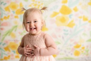 floral milestone session a one year old girl in a Milwaukee photography studio wearing a blush pink tutu dress. She's in front of a yellow. blue and pink floral paper backdrop with her hands on her tummy smiling big