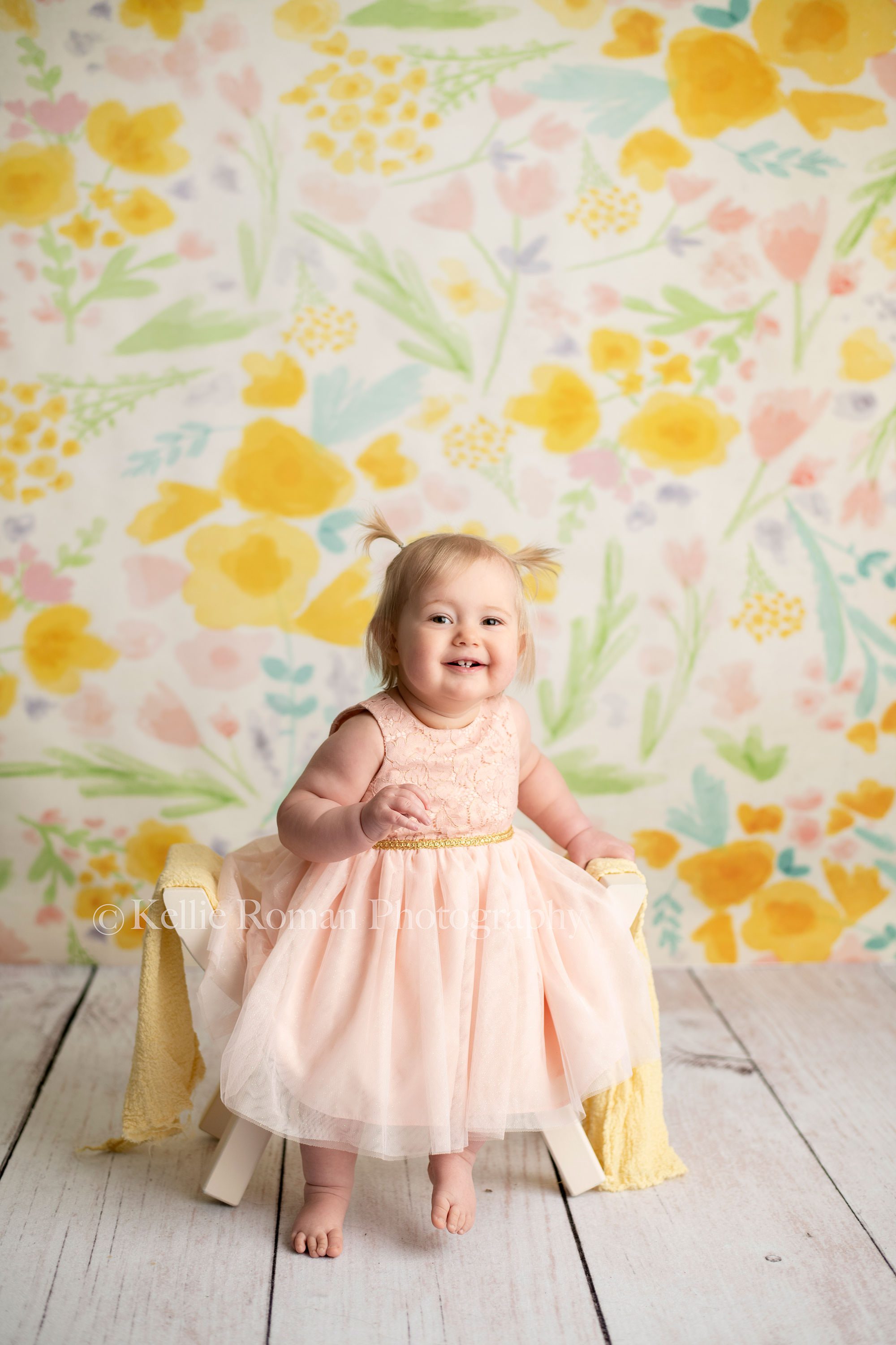 floral milestone session a one year old little girl from kenosha in a Milwaukee photographers studio for milestone session. She is wearing a blush pink dress, and sitting on a white curved bench with a yellow wrap over it. She's in front of a floral backdrop with a white wood floor smiling very big at the camera she has pigtails