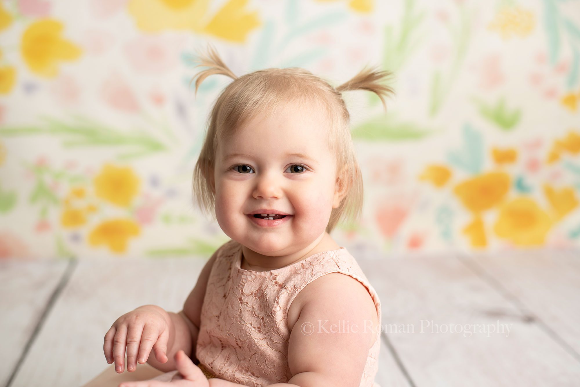 floral milestone session a one year old little girl from kenosha wearing a pink dress in front of a yellow blue pink and purple floral backdrop she's in a Milwaukee photographers studio she is looking at the camera and has a big smile with a few little teeth