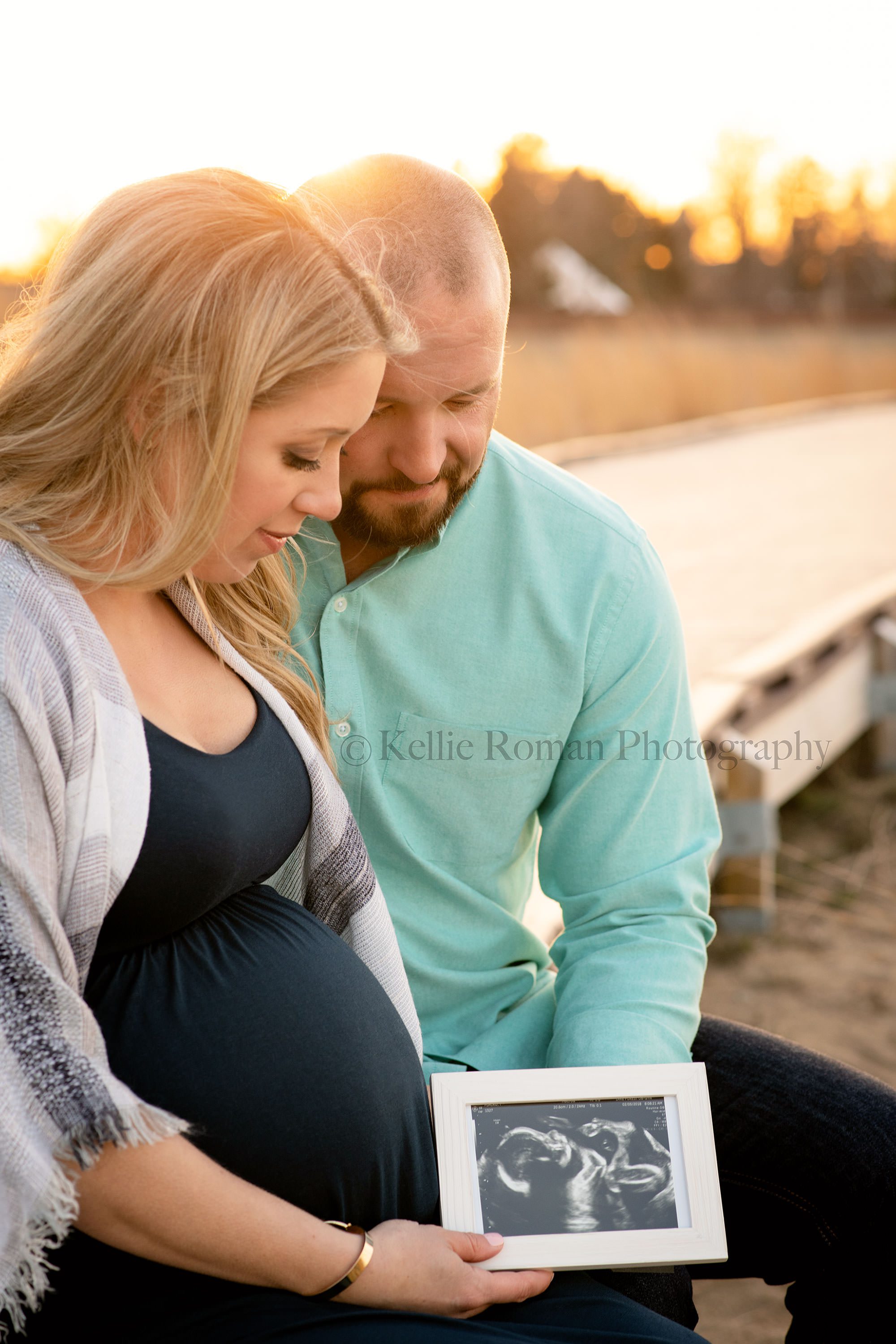 cold night at the beach a man and women husband and wife who are expecting their first newborn from Illinois are on a kenosha Wisconsin beach sitting on a wood walkway. They are holding a framed photo of an ultrasound pictures and looking down at it. The sunlight is behind them while on the beach