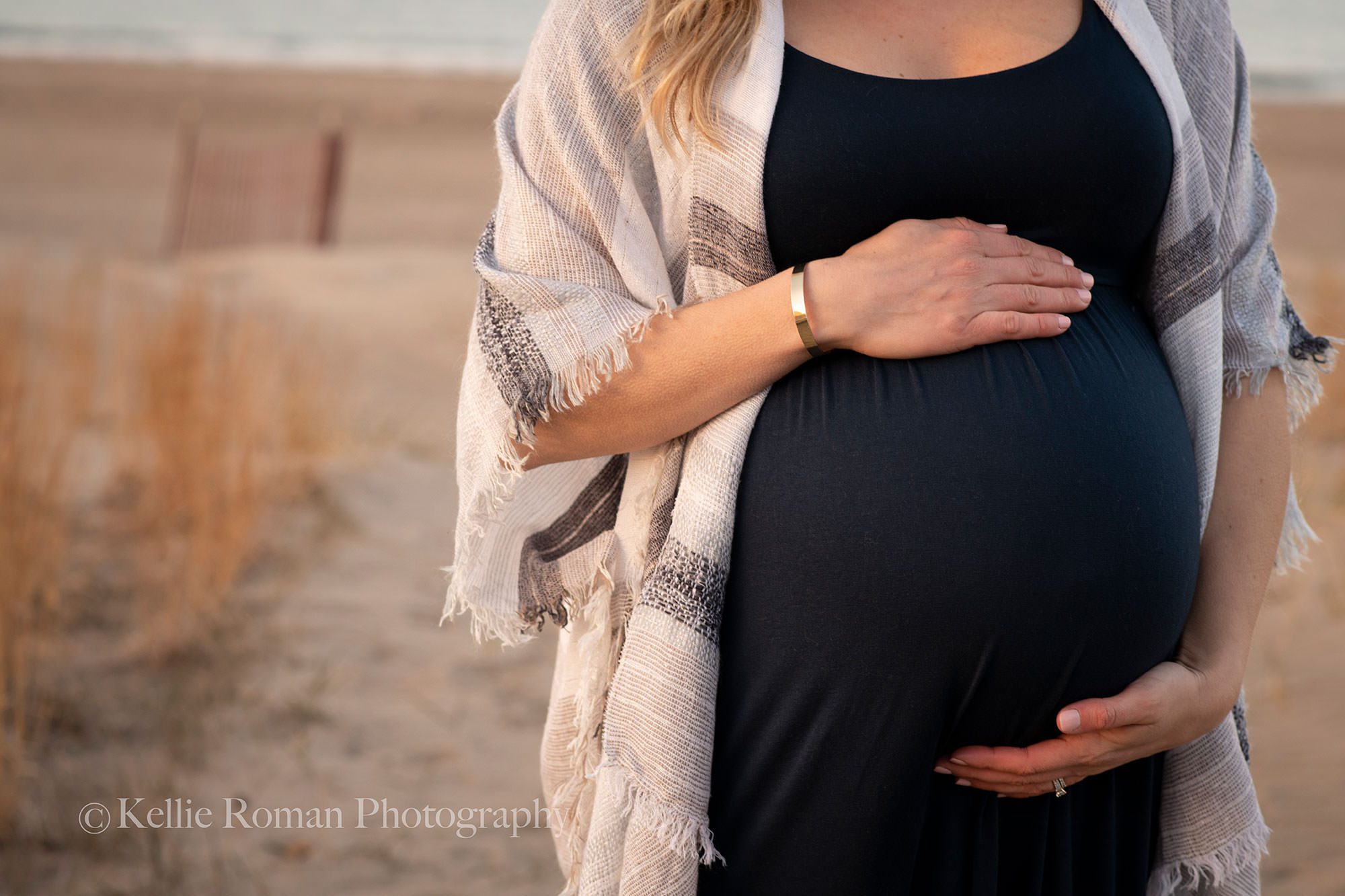 cold night at the beach a women from Illinois on a beach in kenosha who is pregnant holding onto her baby bump. Having her maternity session photographed and wearing a blue dress with sweater over the top sand in the backdrop