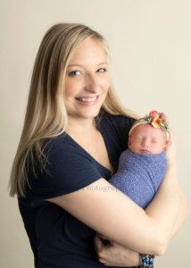 sweet little sister a mother wearing a blue shirt who's from racine Wisconsin is holding newborn daughter in a Milwaukee photographers studio newborn girl is swaddled in a blue wrap and is wearing a blue headband the mother is looking into the camera