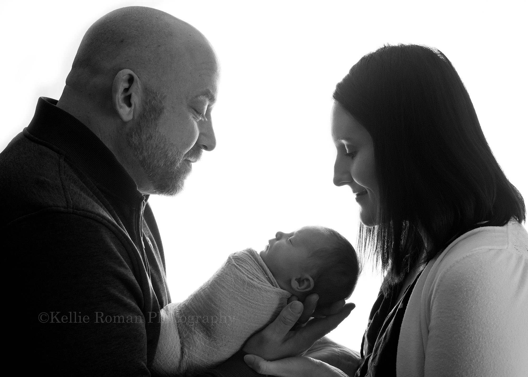 grandmas bunny parents from kenosha with the newborn baby boy in a Milwaukee photography studio. Black and white image that is backlit in studio so it's nice and bright white. father is holding wrapped newborn son and both mother and father are looking down at sleeping newborn