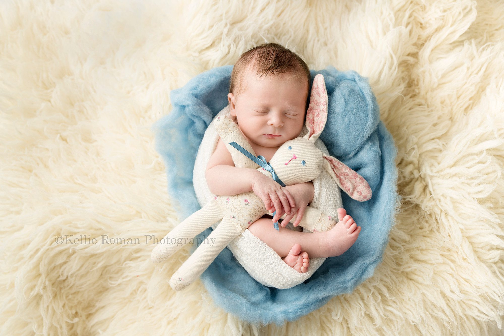 grandmas bunny a newborn baby boy from kenosha in a Milwaukee photographers studio he's wrapped in an ivory swaddle curled up in a posed on top of blue and ivory fluff and and flokati rug he's sleeping and has in grandmas craft bunny tucked into his newborn hands