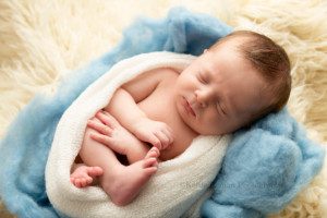 grandmas bunny newborn baby boy from kenosha in Milwaukee photographer studio he's wrapped in ivory swaddler onto of blue and ivory fluff sleeping with feet and hands sticking out of wrap
