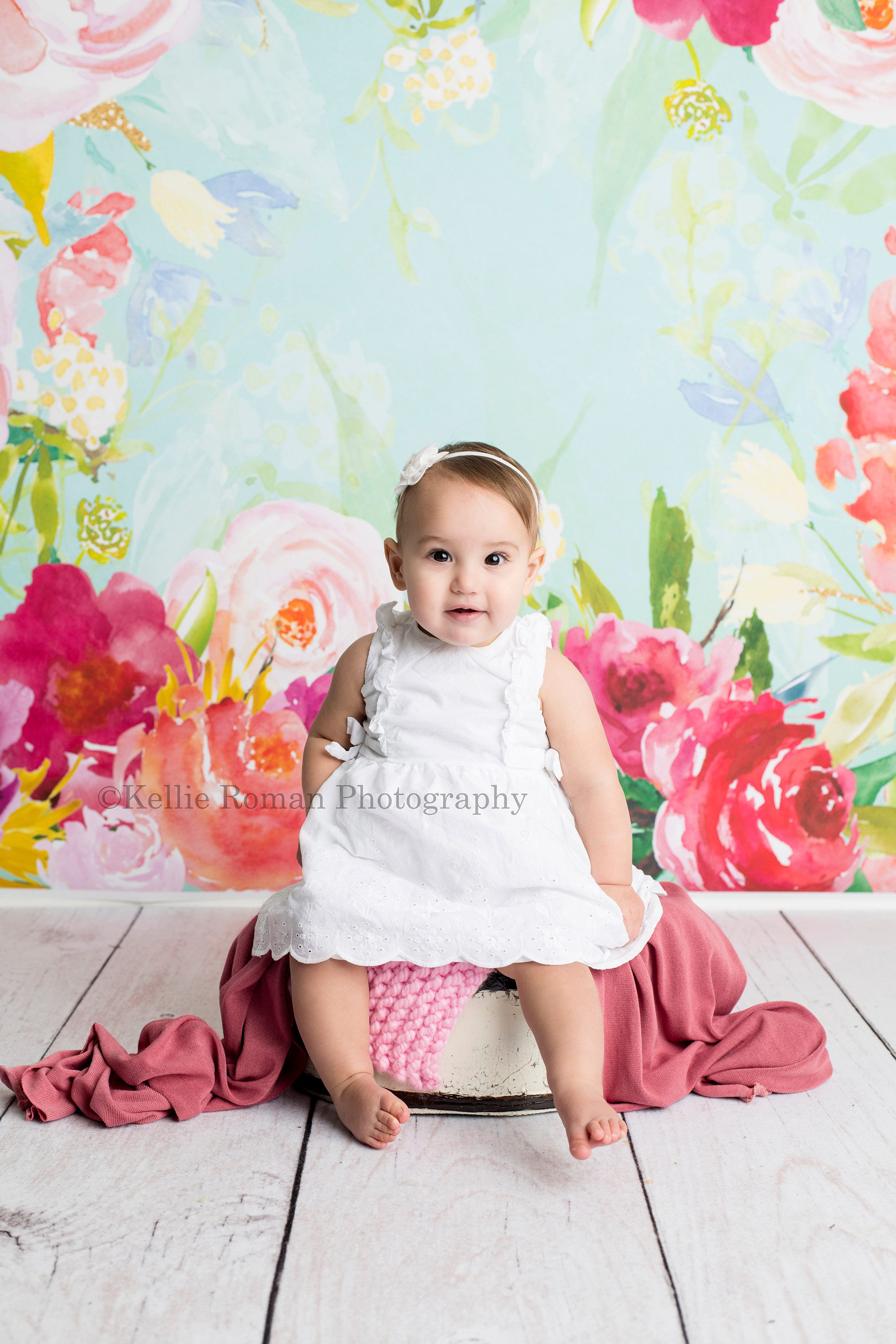 dig in a on year old girl from waukesha in a Milwaukee photographer studio having her milestone and cake smash photos taken. She's sitting on a white tub with a white dress on in front of a floral backdrop