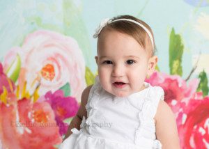 dig in a one year old little girl from waukesha in Milwaukee photography studio wearing a white dress in front of a floral backdrop. She's having her milestone session photographed