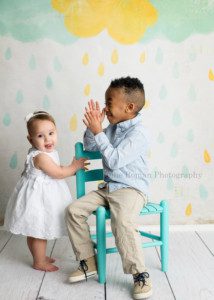 dig in a brother and sister in Milwaukee photographer studio having photo session. Boy is sitting on a teal chair clapping at his sister and his sister is standing looking at the camera. Milestone session in Milwaukee photographer studio