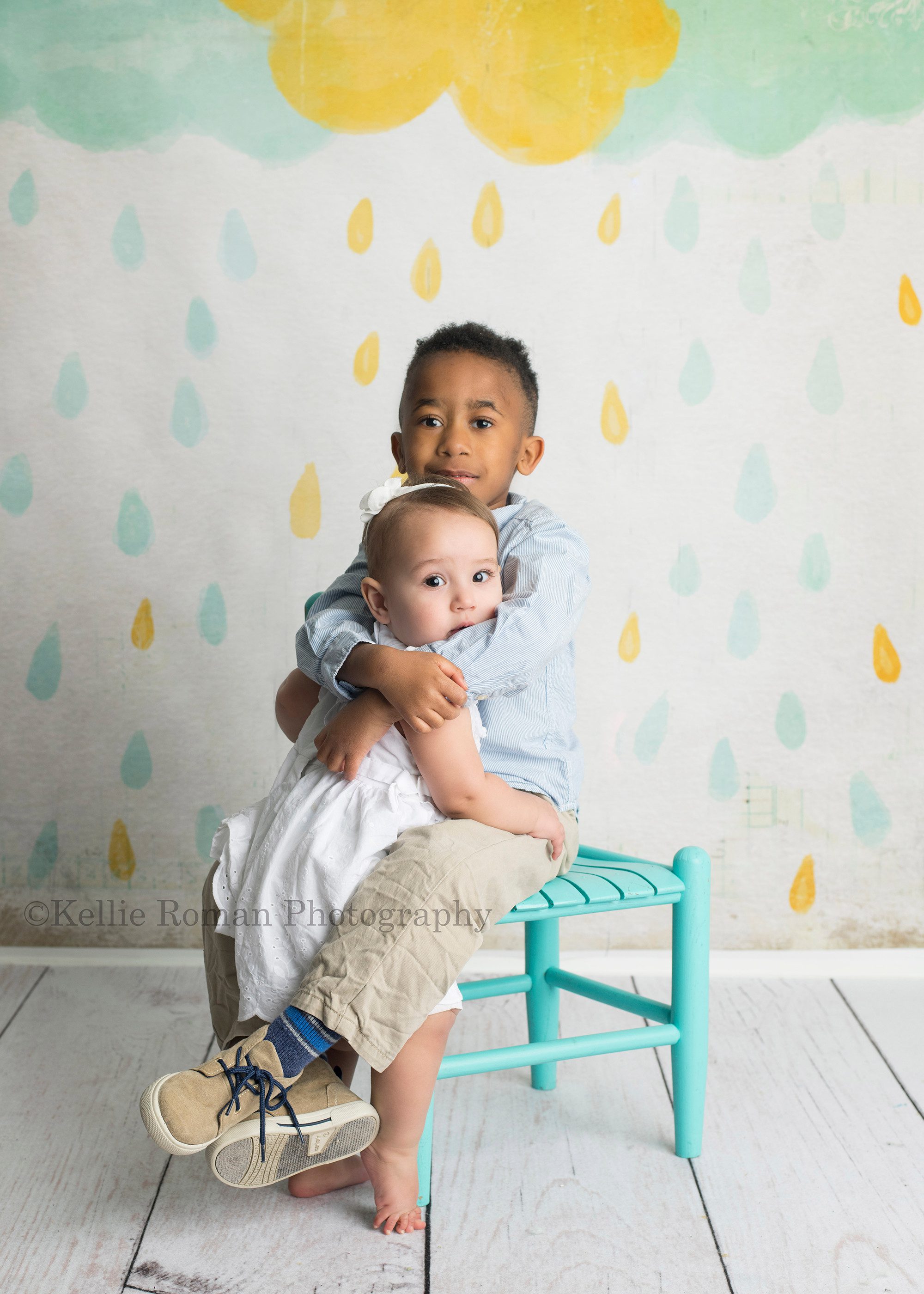 dig in a brother and sister in Milwaukee photographer studio having photo session. boy is sitting on a teal chair and is hugging his little sister from the side. they are in front of a colored cloud backdrop with raindrops.