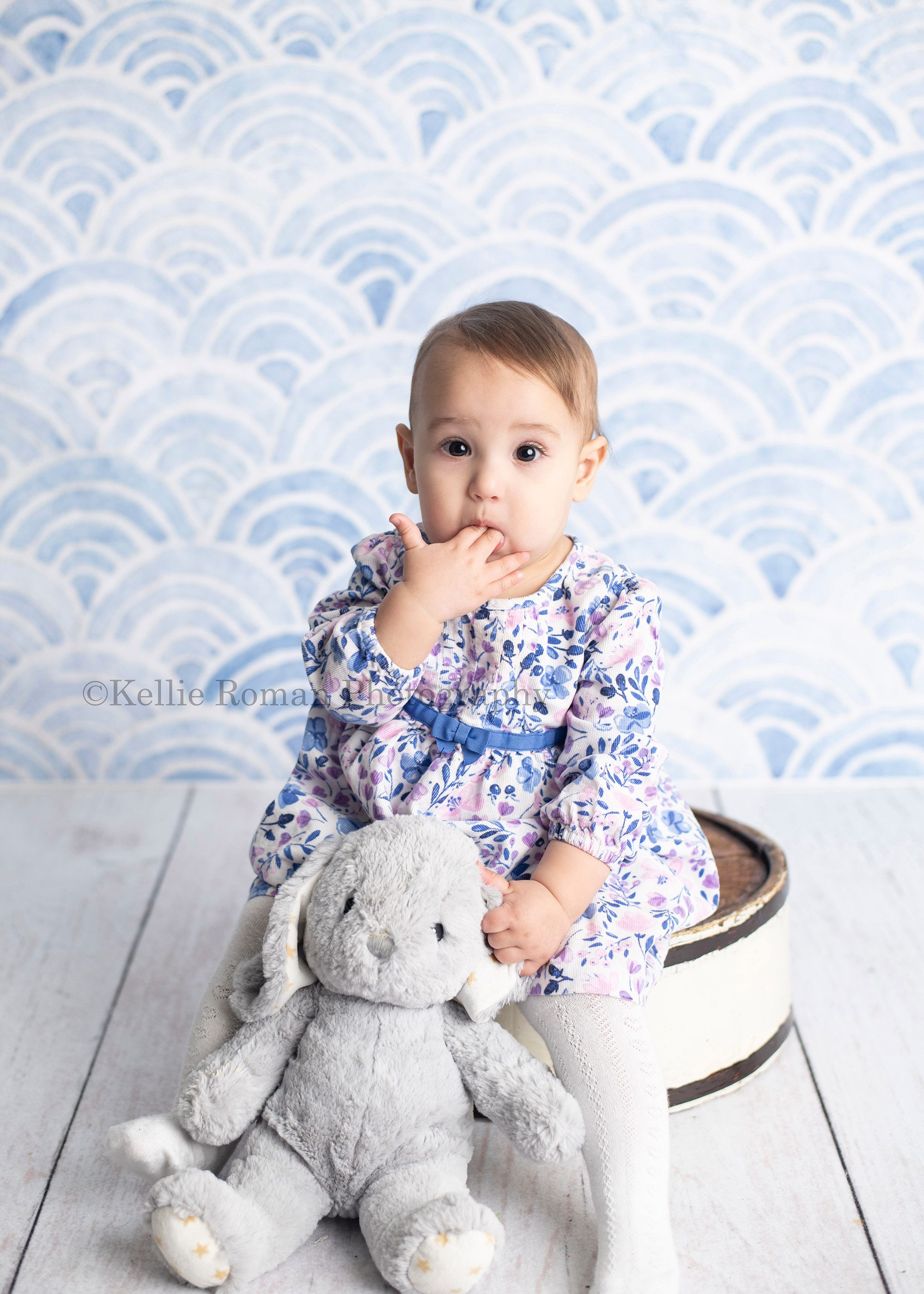 dig in a one year old girl in a Milwaukee photography studio swearing a blue and purple dress sitting on a white tub infant of a blue backdrop. She has her fingers in her mouth and is holding a grey bunny. One year old milestone session