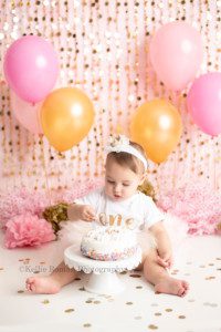dig in a little birthday girl in a Milwaukee photography studio for her milestone cake smash session. She's sitting behind a white cake on a white cake stand surrounded by gold confetti she has a gold crown on with a onesie that says one. Her birthday cake smash theme is pink and gold