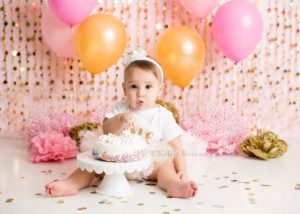 dig in a one year old little girl from waukesha in a Milwaukee photography studio sitting behind a white cake with colored sprinkles on a cake stand with frosting and cake covering her hand and mouth. She's looking right into the. camera, while sitting on the floor surrounded by gold confetti. She has a festive pink and gold birthday theme