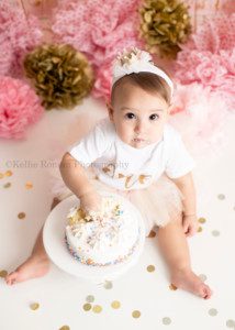 dig in a one year old girl from waukesha in Milwaukee photography studio smashing into a small white cake with colored sprinkles shot from above. She is looking up at camera with frosting covering her lips. There is gold confetti on the floor and a pink and gold festive set up