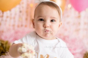 dig in a one year old girl from waukesha in a Milwaukee photography studio with cake all over her hand and face. She's wearing a white onesie that says ONE and the backdrop colors are pink and gold