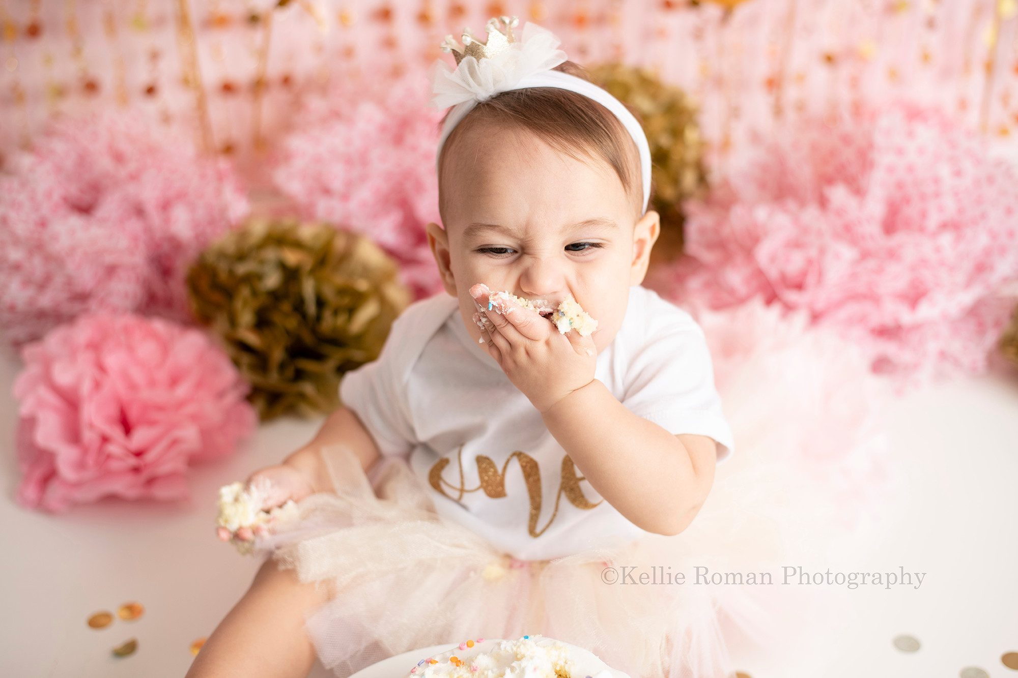 dig in a one year old girl from waukesha in a Milwaukee photography studio smashing cake into her mouth for her one year old milestone photography session. The colors of the session are pink and gold