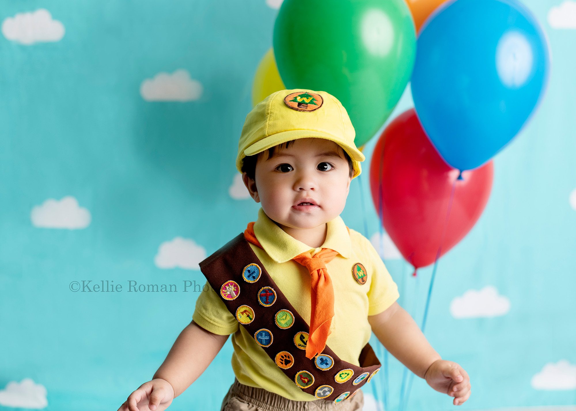 up up and away a kenosha boy in Milwaukee photographer studio wearing a costume that looks like the character in the movie UP. He has a scout uniform one with brown sash and orange scarf. He's infant of a blue cloud backdrop with colored balloons