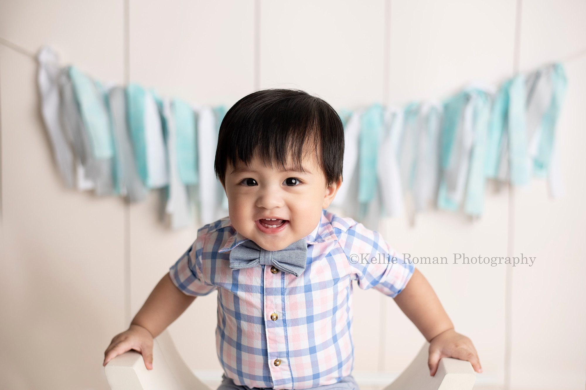 up up and away a one year old boy in Milwaukee photography studio sitting on a white curved bench smiling at the camera close up of his face. He is wearing pink and blue plaid shirt and a grey bowtie. He's in front of a cream wood backdrop with rag banner