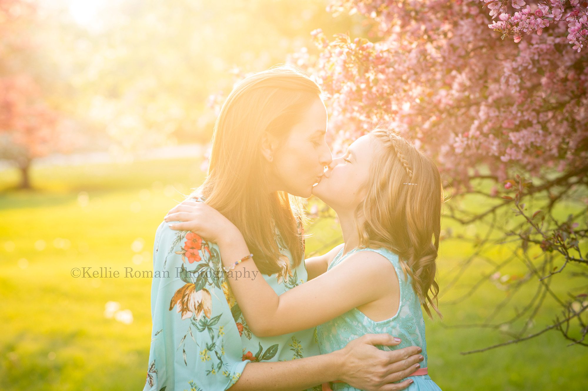 sunlit blossoms a mother and daughter from kenosha standing in a Milwaukee park kissing and hugging each other the sunshine is peaking behind them through blossoms on trees