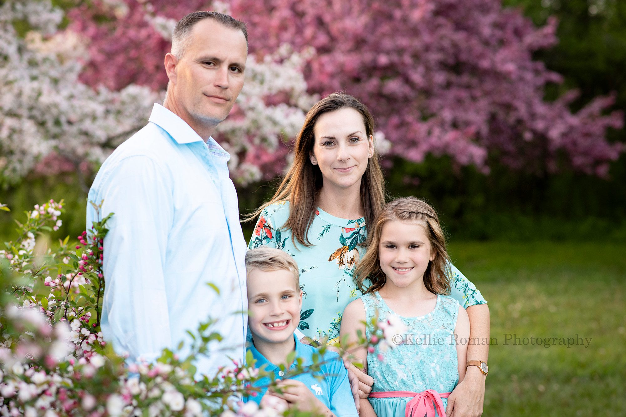 sunlit blossoms a family standing in a park in Milwaukee with crabapple blossoms surrounding them they are all wearing shades of blue clothes and smiling at the camera