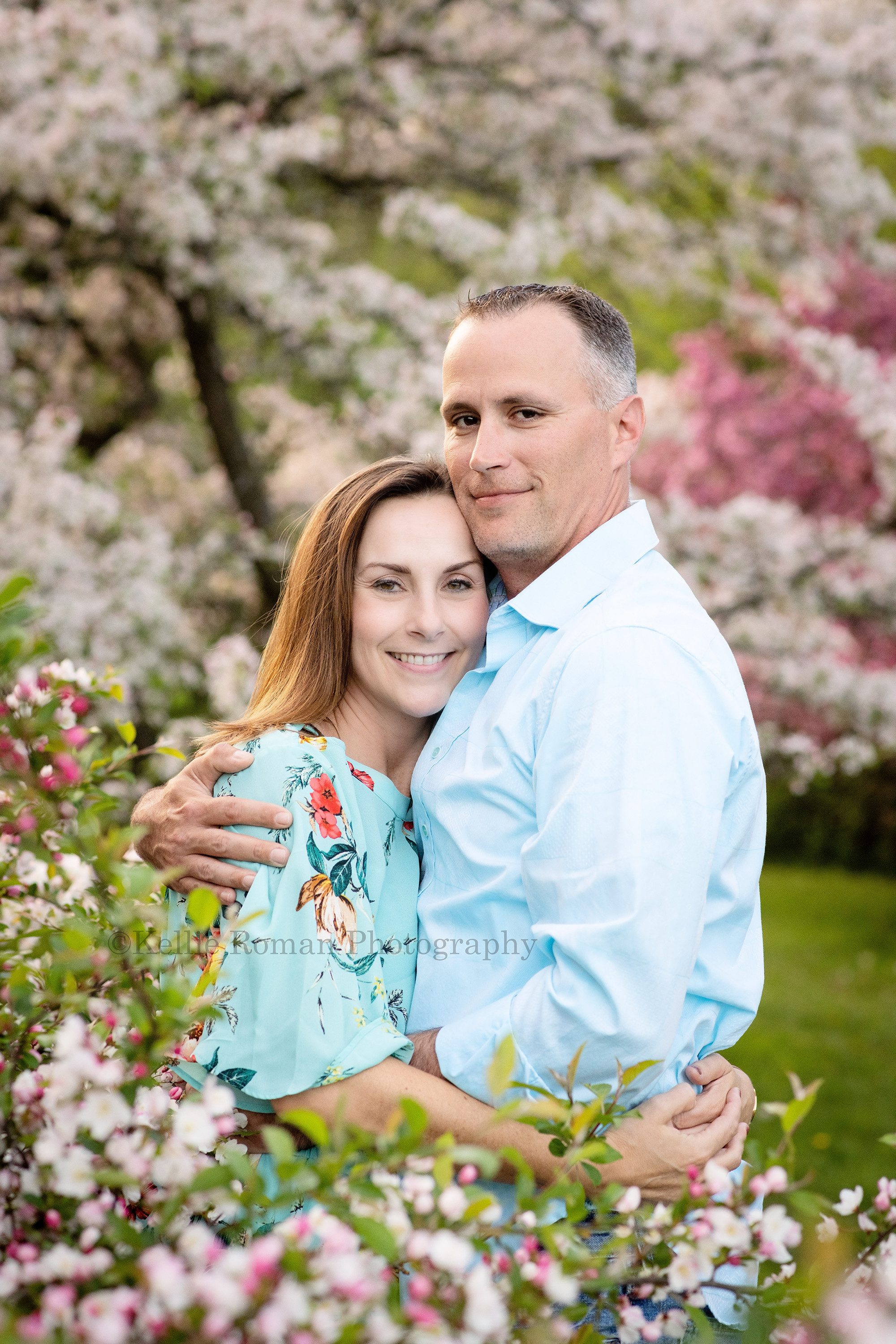 sunlit blossoms married couple standing within trees that are blooming while hugging each other and looking at the camera park within Milwaukee County