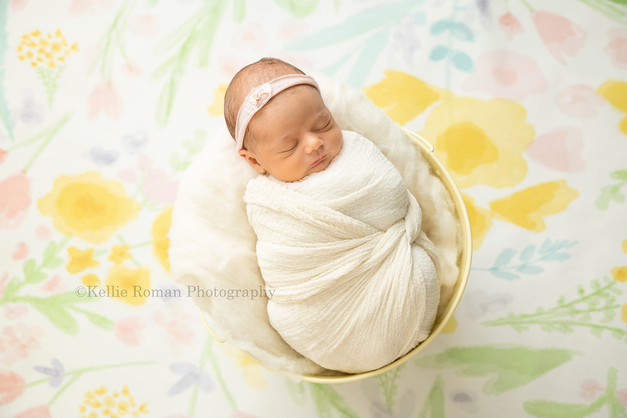 perfect newborn girl a baby newborn girl wrapped in an ivory swaddle fabric laying in a yellow metal bucket she has a headband on and the bucket she's in is onto of a floral backdrop her parents are from Madison Wisconsin