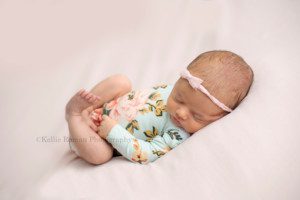 perfect newborn girl a baby girl laying on her back in a posed position so her feet are curled up she's sleeping and has on a blue and pink floral romper with a very light pink bow she's photographed in a milwaukee photographer studio