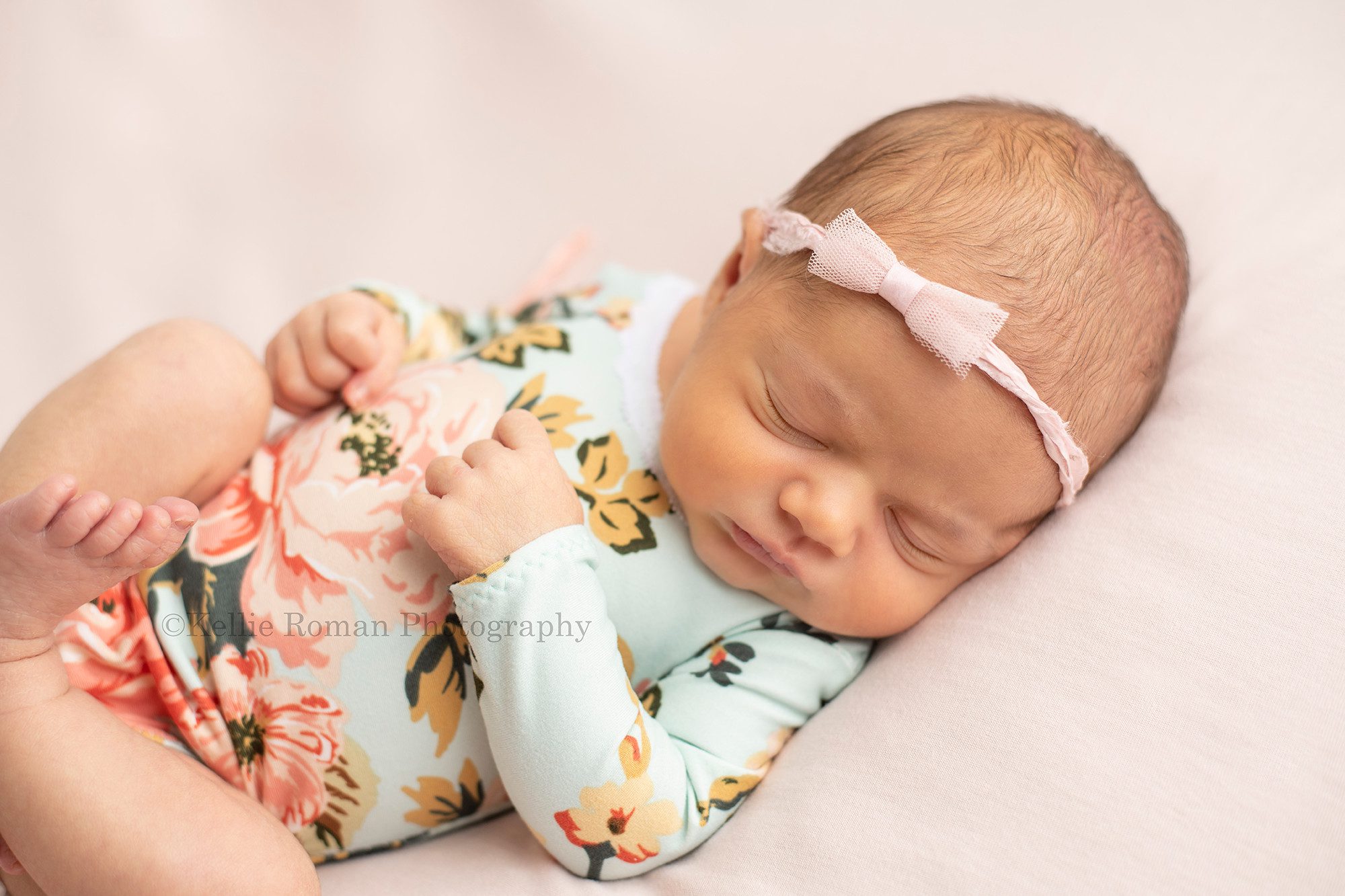 perfect newborn girl a little newborn girl wearing a floral blue and pink romper is laying on her back sleeping on top of a pink blanket she has a very small pink bow headband on she's being photographed in a milwaukee photographers studio