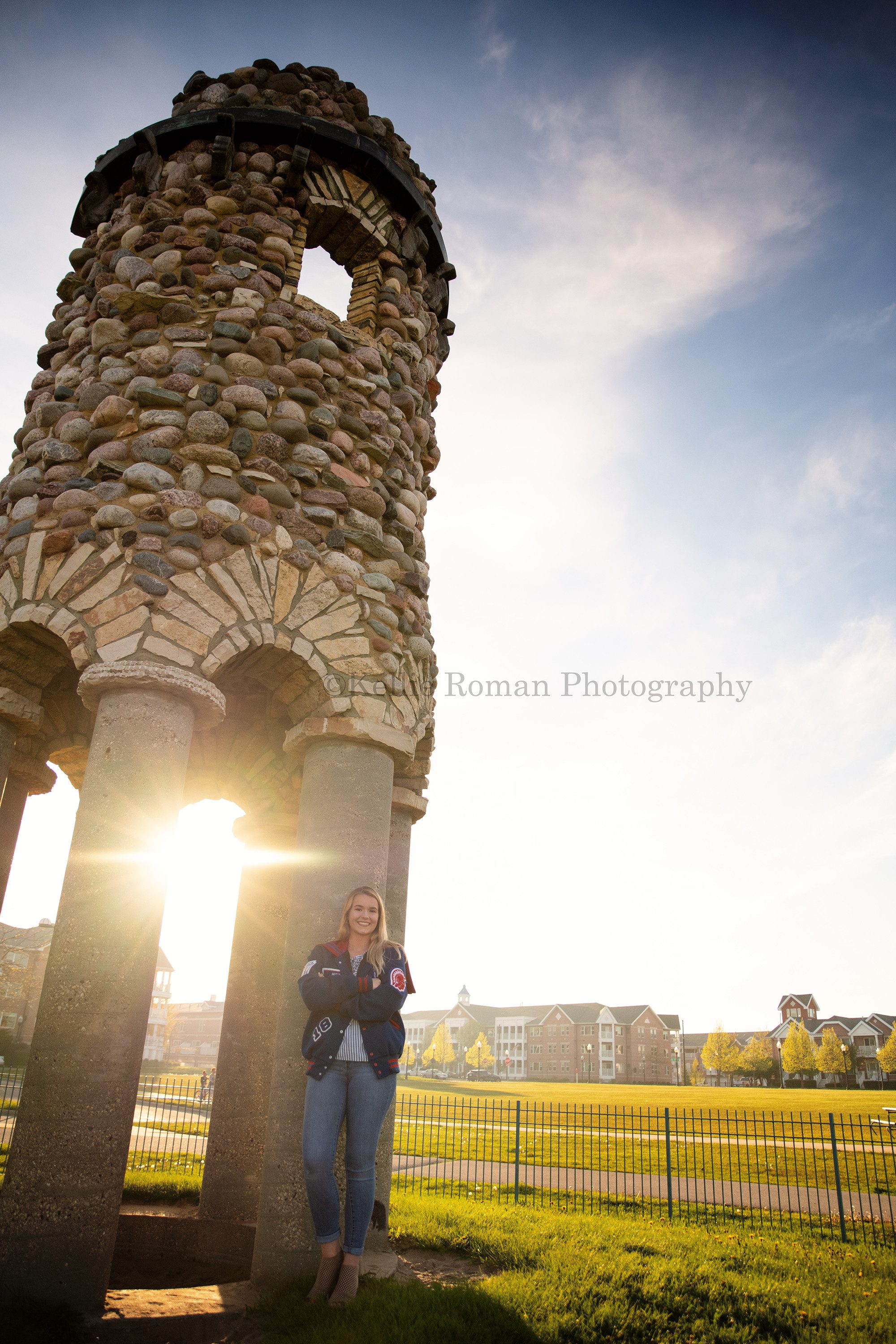 senior beach session a girl from kenosha who is a high school senior is standing in front of a brick and rock tower at kenosha marina she is wearing her letterman jacket and the sun is shining from behind her she's being photographed by a kenosha photographer