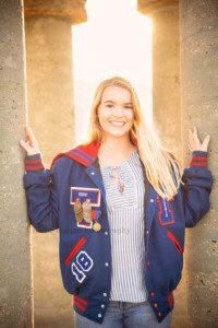 senior beach session a girl is standing with cement pillars at a marina in kenosha the sun is glowing from behind her through the pillars she is wearing her blue and red letterman jacket and is smiling at the camera