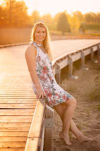 senior beach session a high school senior girl from kenosha on a kenosha beach sitting on a wood boardwalk she is wearing a floral dress with no shoes and is looking at the camera the gold sun is coming from behind her and is glowing through her hair