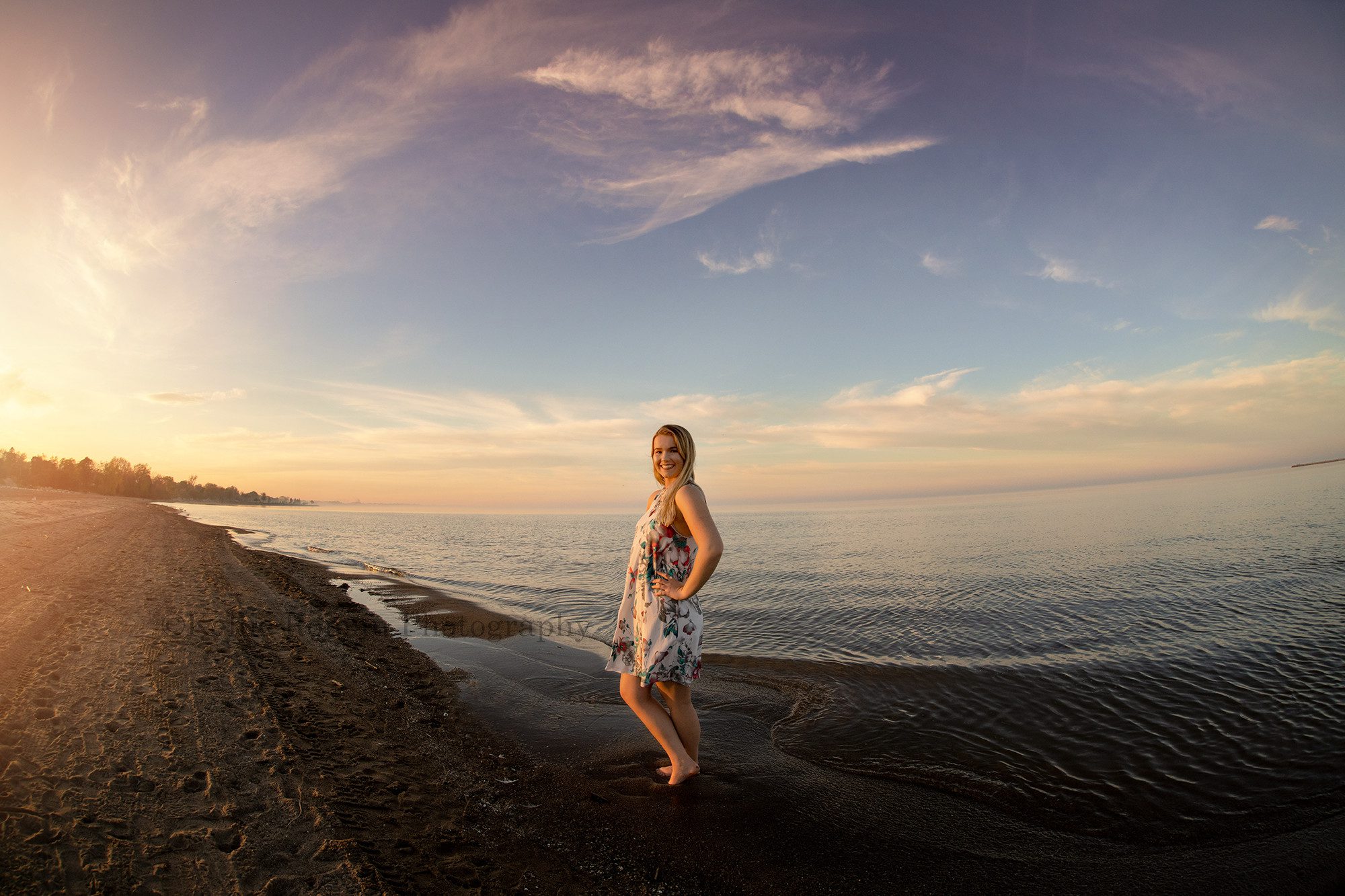 senior beach session a high school senior girl from kenosha Wisconsin is standing on a beach on Lake Michigan she's wearing a dress with flowers and the sky behind her is lit up with pastel colors a photographer from milwaukee is photographing her