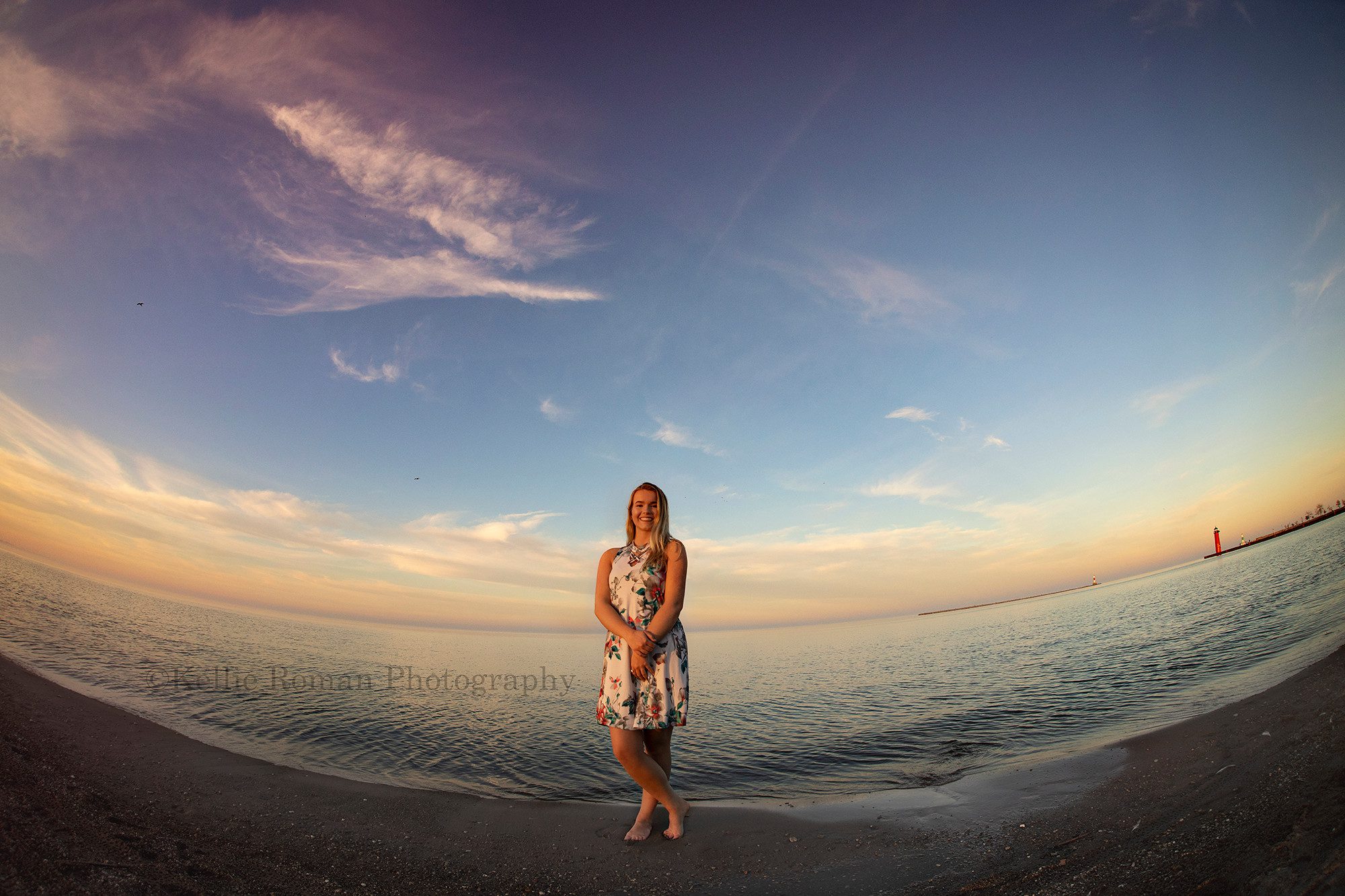 senior beach session a high school senior from kenosha Wisconsin is being photographed on the beach she is standing in the sand right in front of the water the shot was taken with a fisheye lens and the sky is shades of orange and pink the red Simmons light house is seen in the background