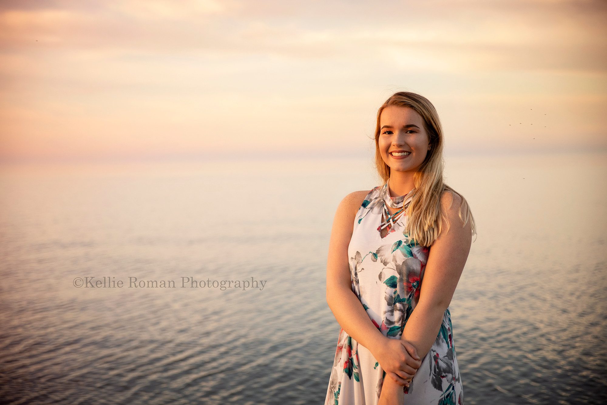 senior beach session a high school senior from kenosha Wisconsin is on the beach in front of lake Michigan water she is wearing a floral dress and the lake water and sky is lit up pastel colors