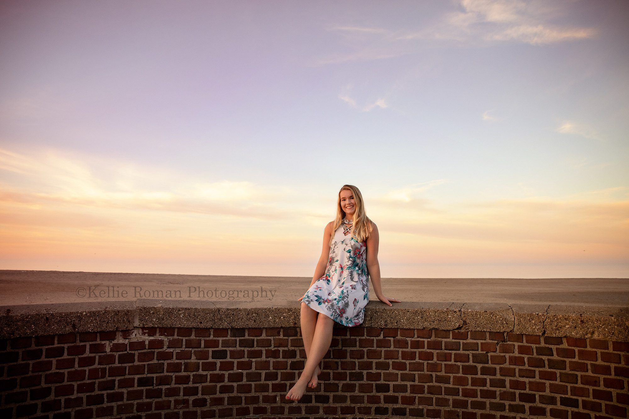 senior beach session a young high school student is sitting on a brick wall at Simmons beach in kenosha she is barefoot and wearing a floral dress she's smiling into the camera and the sky behind her is shades of pink and orange