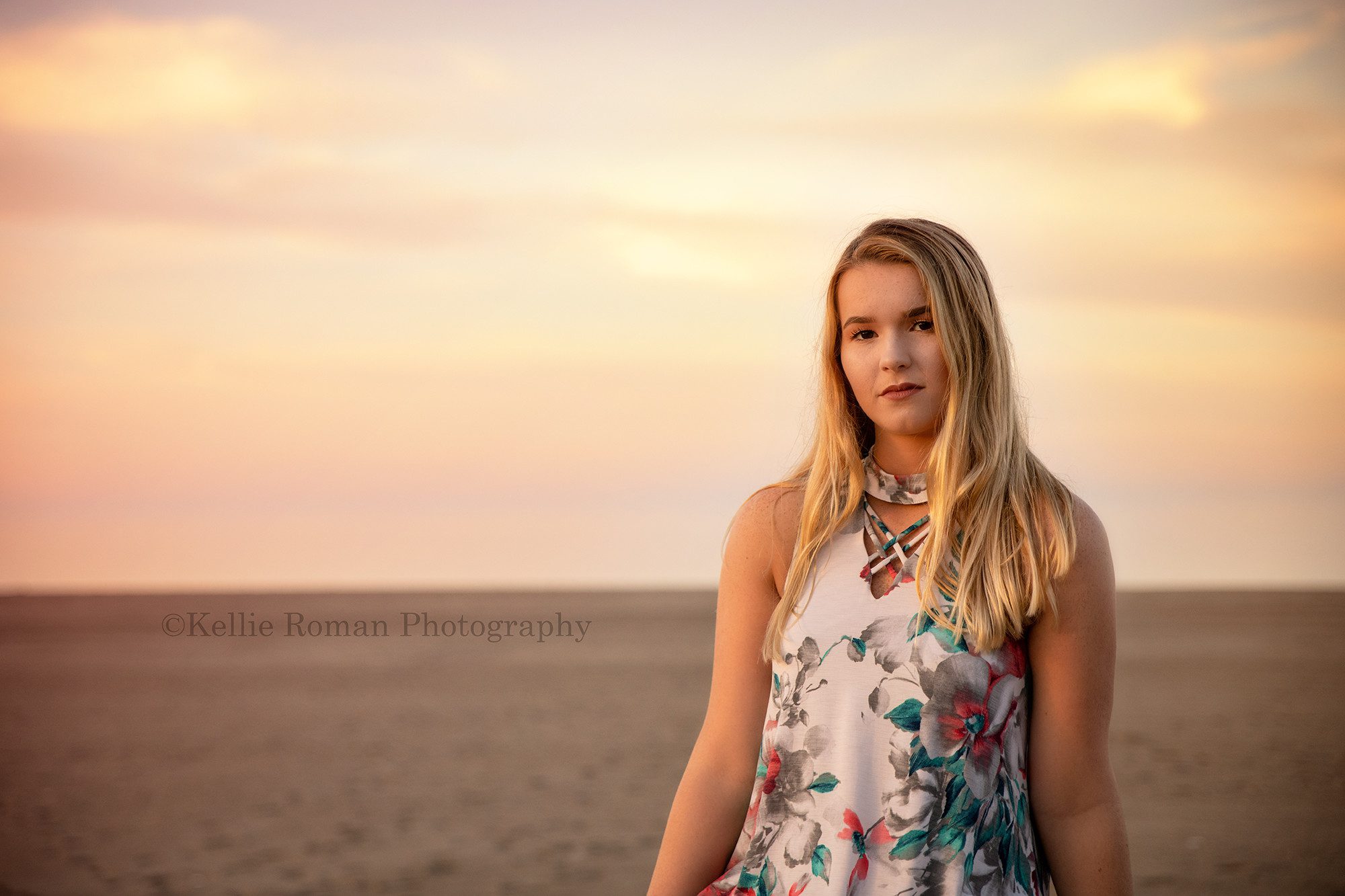 senior beach session a high school senior girl is sitting on a brick wall at the beach in kenosha Wisconsin she has a floral dress on and long blonde hair she's looking seriously into the camera the sky behind her is shades of pastel