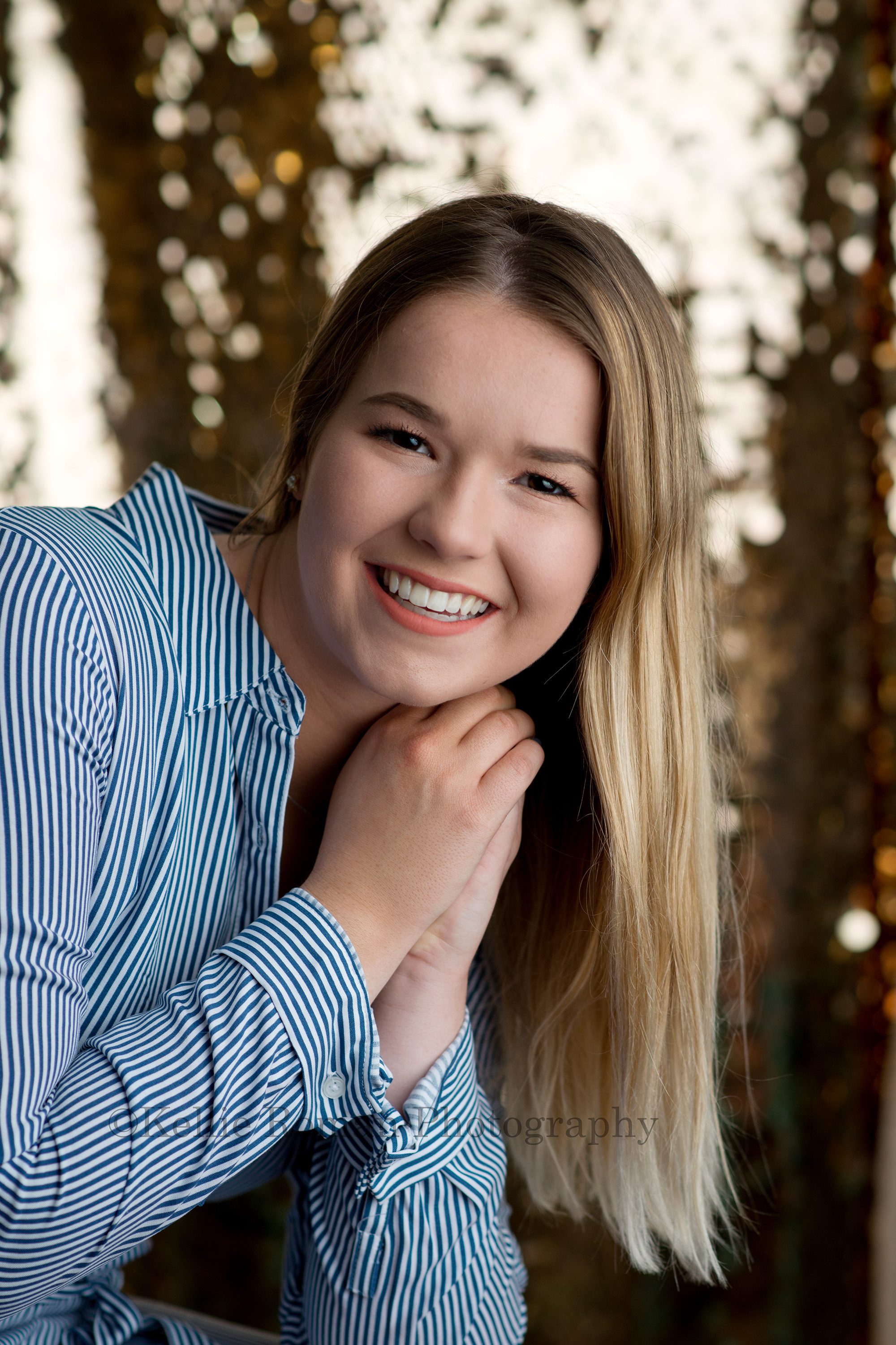 senior beach session a high school senior from kenosha is in a Milwaukee photographers studio she is wearing a blue and white striped dress and has her hands by her face looking and smiling into the camera she is in front of a gold sequin backdrop