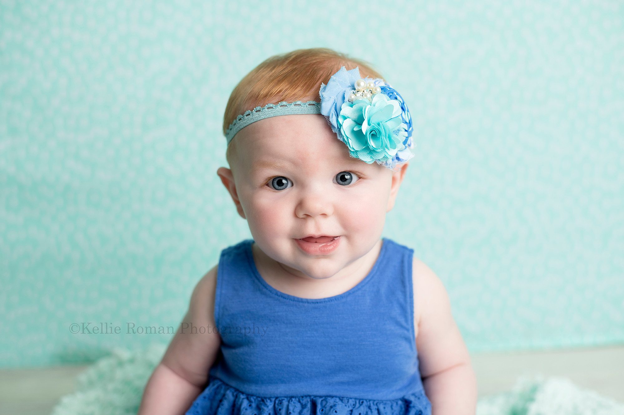 six month session a six month old infant girl is wearing a royal blue dress with matching headband she's sitting on a mint green blanket in front of a mint green backdrop in a milwaukee photographers studio she's smiling at the camera