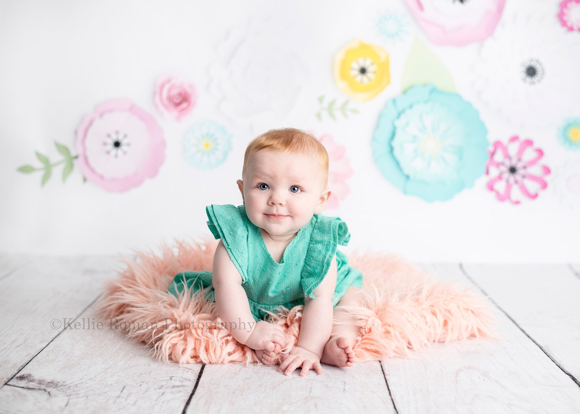 six month session a six month old infant girl is sitting on peach fur with a white backdrop with colored flowers and a white wood floor she has a green dress on and has a sweet innocent expression on her face while looking up with big blue eyes
