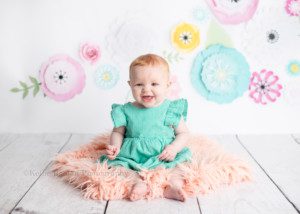 six month session a little red headed girl is in a milwaukee photography studio she's sitting on a beach fur rug onto of a white wood floor the backdrop is white with colored flowers the little girl has a green dress on and is smiling