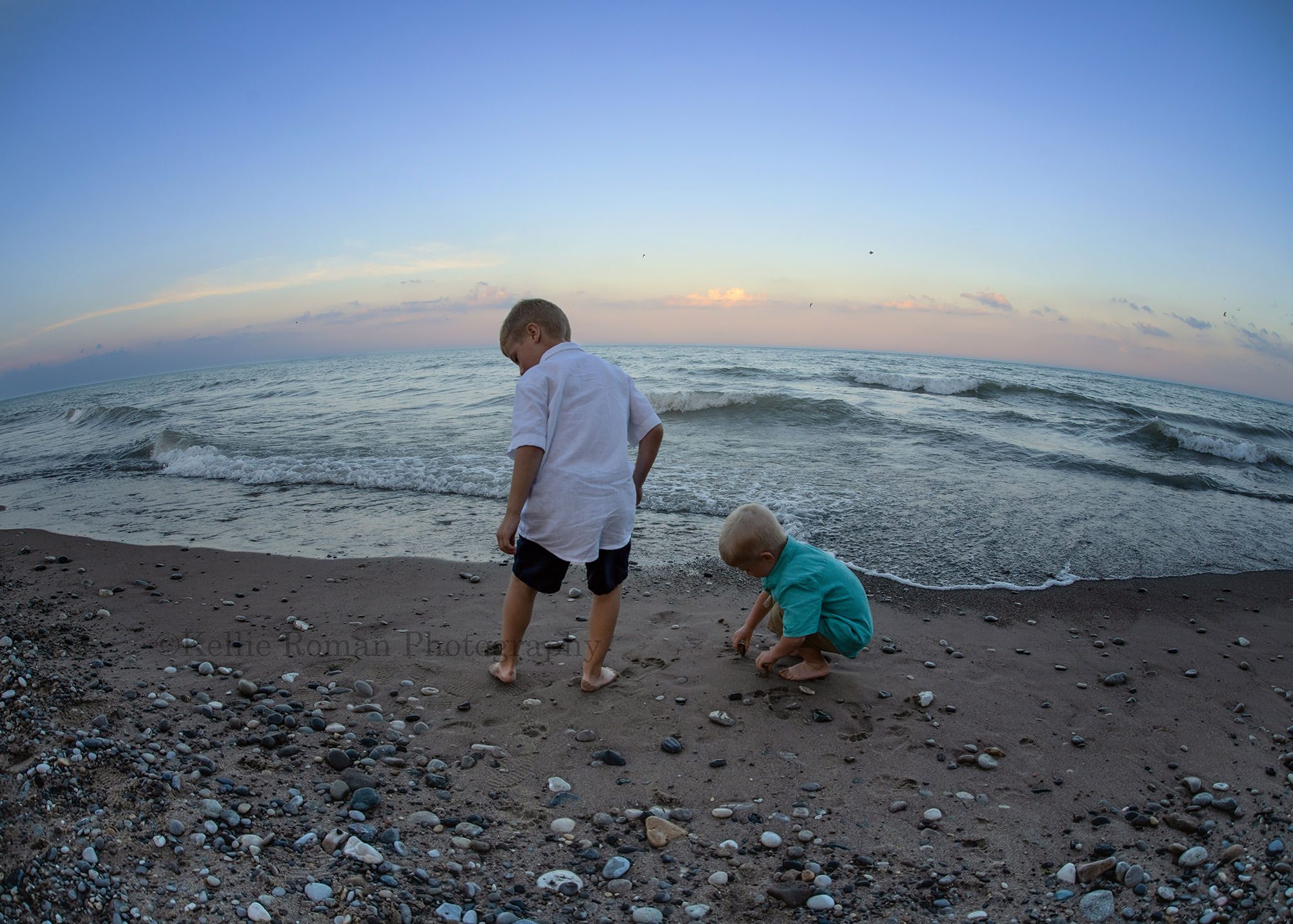 racine family session two brothers on a rocky sandy beach in racine Wisconsin they are picking up rocks and looking down the sky has shades of pastels and is during the sunset