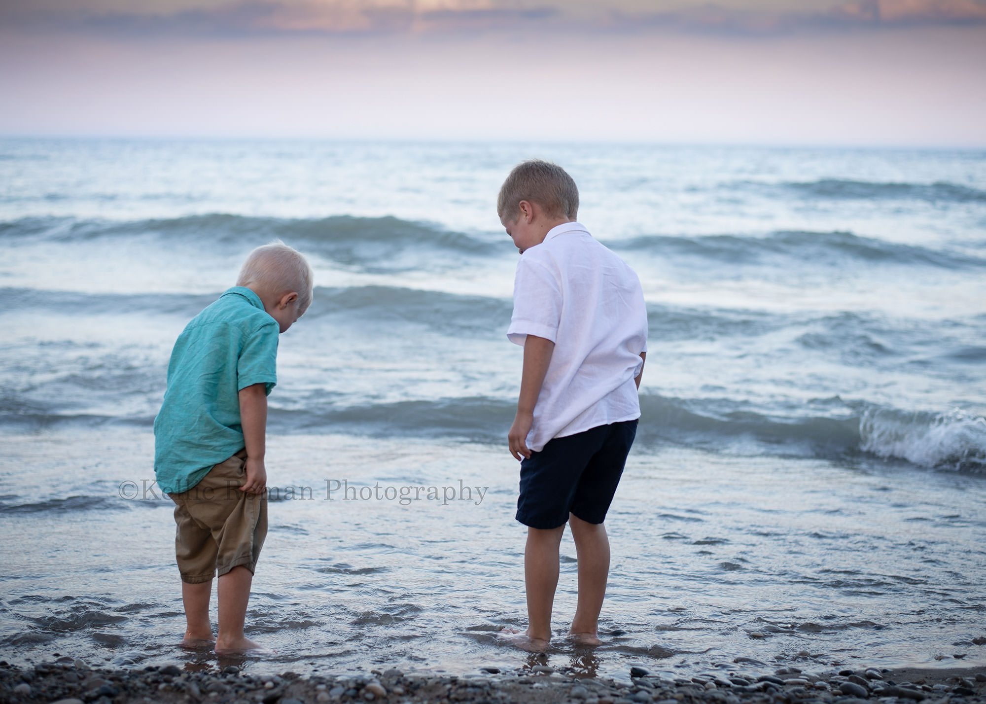 racine family session two brothers looking down on a beach in racine they have their feet in the water on a rocky beach they have on shorts and button up shirts the sky is shades of pastel with some clouds