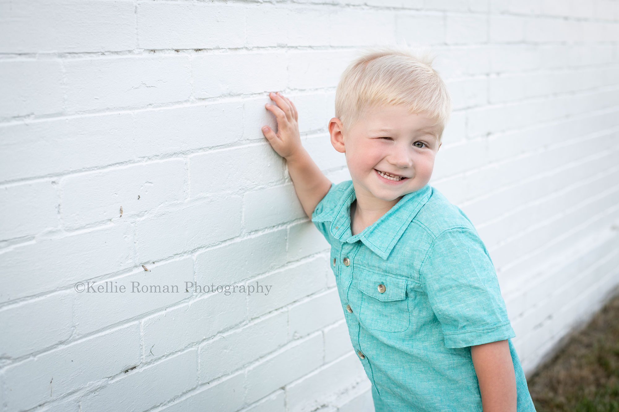 racine family session a little boy wearing a teal button up shirt is standing in front of a white brick building near a beach in Wisconsin he's winking at the camera