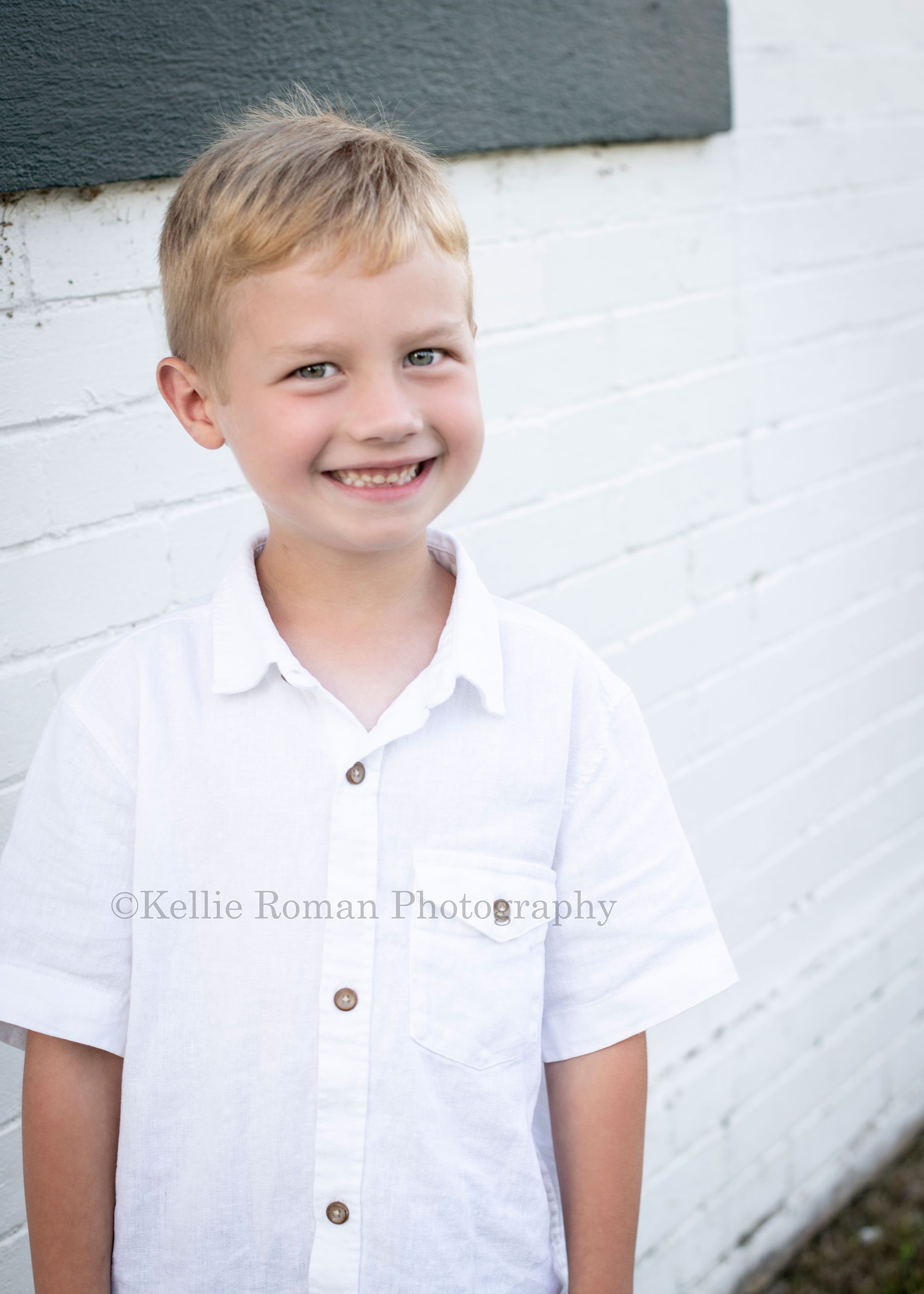 racine family session a young boy wearing a button up white shirt is standing in front of white brick building he is standing and smiling at the camera