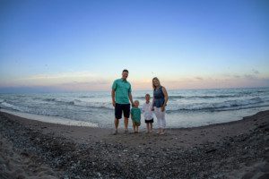 racine family session a family on a beach in racine who is standing in the water of Lake Michigan parents and two boys are looking at the camera white the sky is shades of pastels the sand is full of large rocks