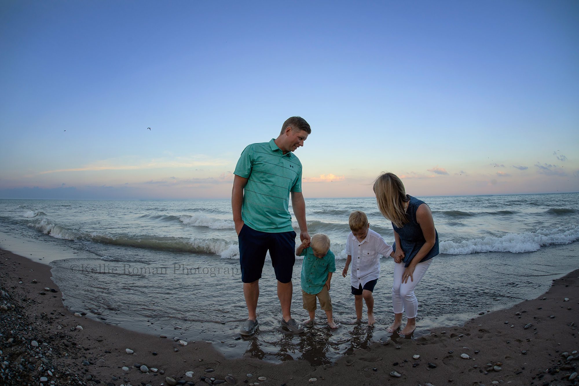 racine family session a family from racine standing in the water of Lake Michigan in racine Wisconsin dad is on one end with two boys in between and mom and on the other end they are looking down at their feet as the water washes in they have on shorts and button up shirts the sky is shades of pastel since the sun is setting bird are flying in the background